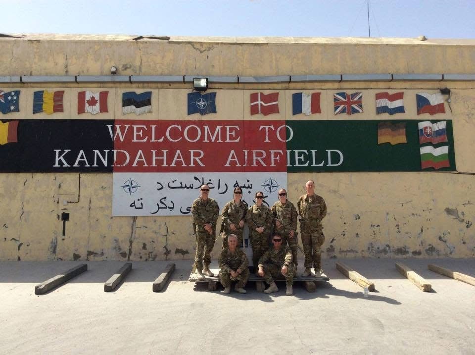 A group of seven people in camouflage uniforms in front of a sign reading "Welcome to Kandahar Airfield"
