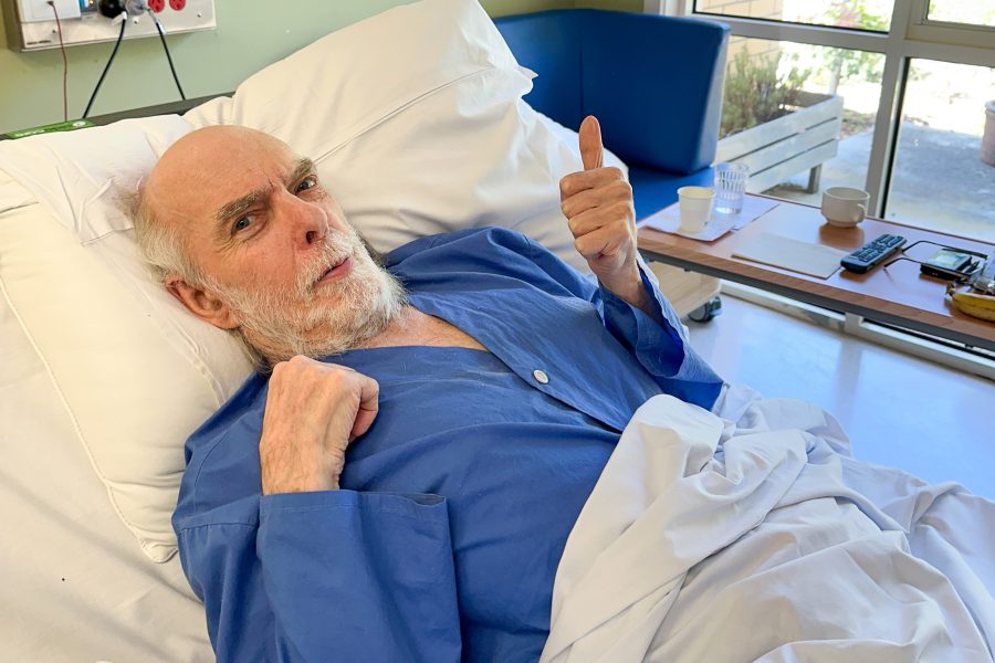 Len Harrison lays in hospital bed wearing blue robe and displaying a thumbs up
