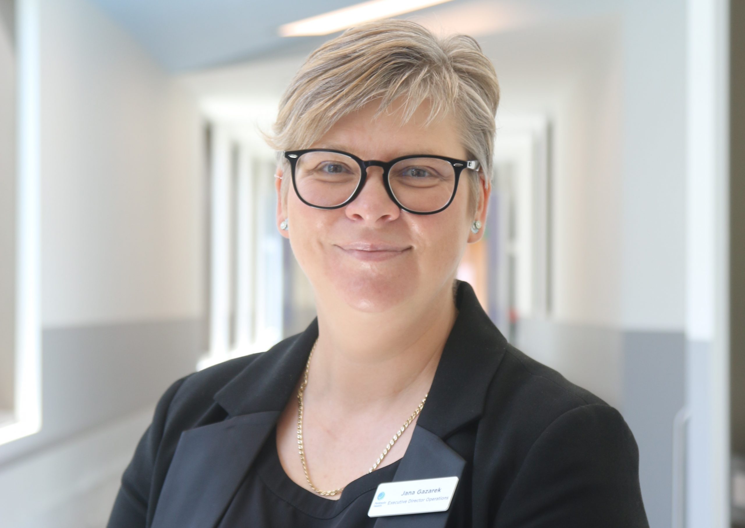 A blonde woman with glasses stands in the corridor of Frankston Hospital smiling